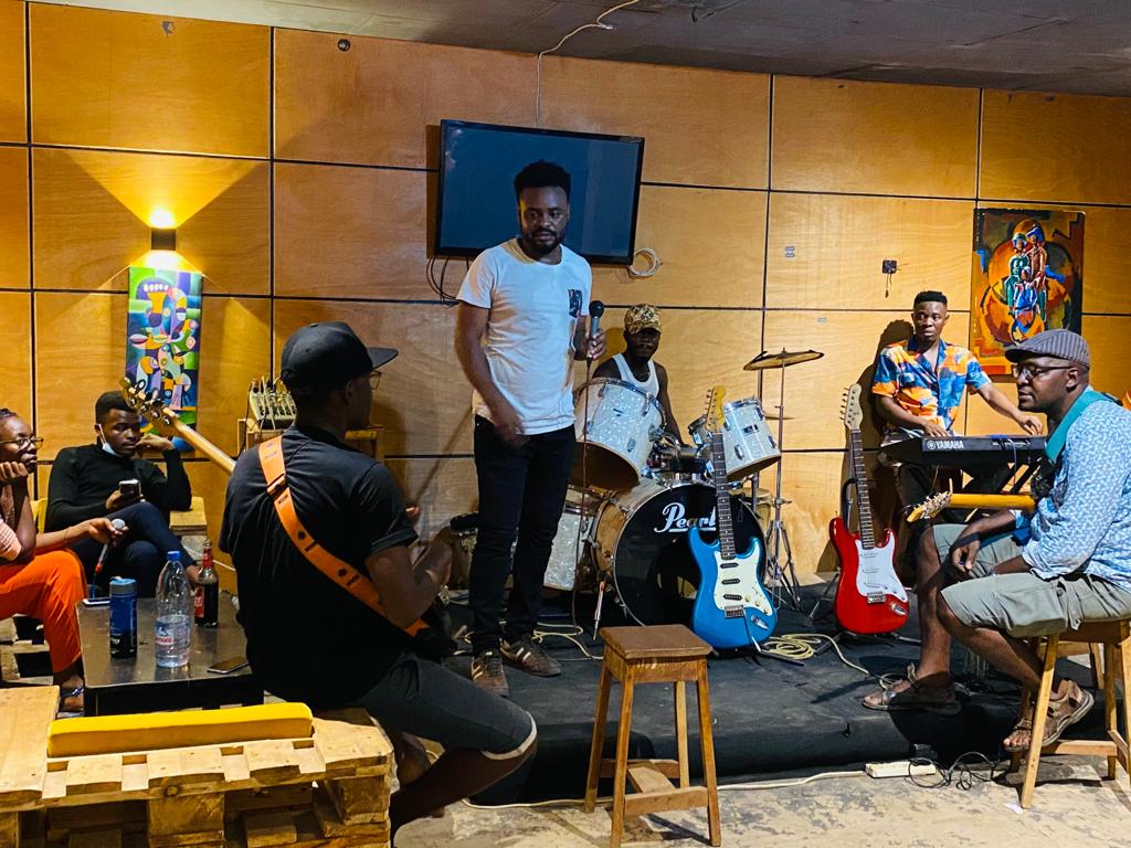 A group of five people sitting and standing in a recording studio, playing electric guitar, drums and electronic keyboard. Two other people are sitting to the left.