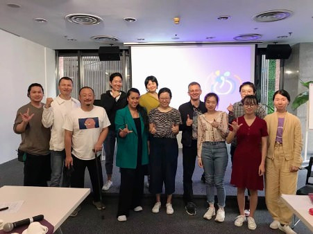 12 people standing together in a group, signing a positive message with their hands.